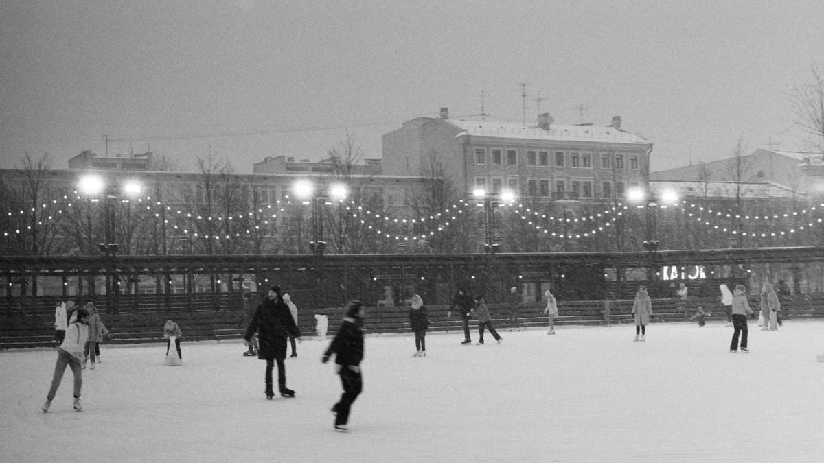Black and white photo of people ice skating outdoors at night, under string lights in a winter setting.